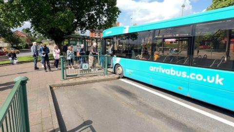 A single-decker turquoise bus parked in an open-air bus stand with a handful of passenger queuing to board. Green railing and a small bus stop can be seen alongside the bus.