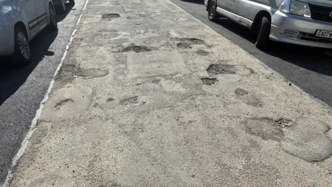 Road surface rough in the middle but resurfaced at the very sides, next to parked cars