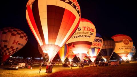 A number of hot air balloons illuminated at night time. They are stationary on a field, with crowds of people looking on.