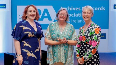 Three people stood in front of a screen at the awards ceremony. Catherine is stood at the centre and is holding an award, with two other people either side.