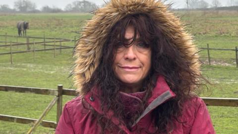 Jann Turner who is standing outside wearing a dark pink jacket with its fur-edged hood up over her head. She has long dark hair. Behind her are fenced fields and in the distance on the left is a horse.