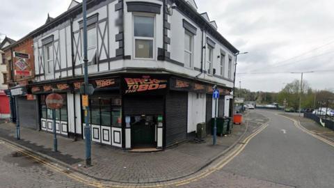 A bar on a street corner with the a 'Back to the 80s' sign overhanging the eaves. The two-storey end terrace building has a black and white painted facade and can be seen with a shutter drawn over the door. 