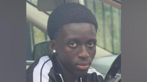 Baye Bireme Fall - a boy with short black hair, wearing a black and white striped tracksuit top, looking at the camera. He is sitting in a car.