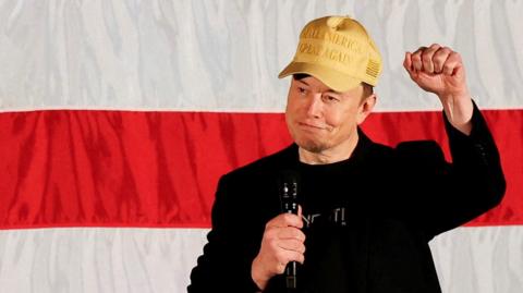 Elon Musk holding a fist up wearing a 'Make America great again' hat