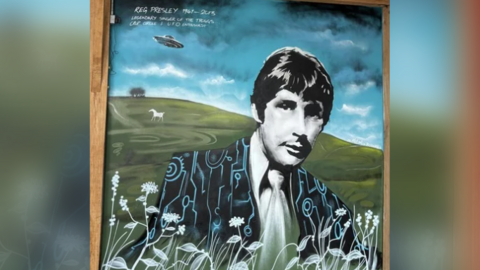 A spray-painted and acrylics portrait of Reg Presley wearing a black suit covered in crop circles, with Alton Barnes White Horse and a UFO in the background.