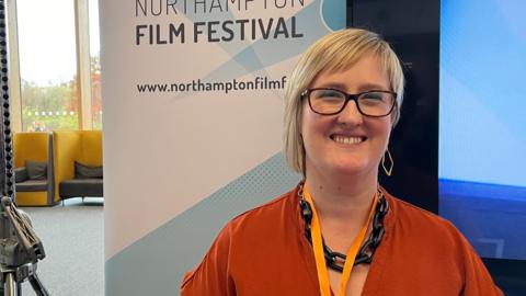 Becky Carrier, who has short blonde hair and wears glasses stands in front of a Northampton Film Festival banner. She is in an orange coloured blouse with a V-neck and is wearing thick, black chain style necklace and a light orange lanyard.
