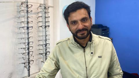 Tipu Sultan, an optical assistant, at his shop. He is wearing a green jacket and there is a glasses stand behind him. 