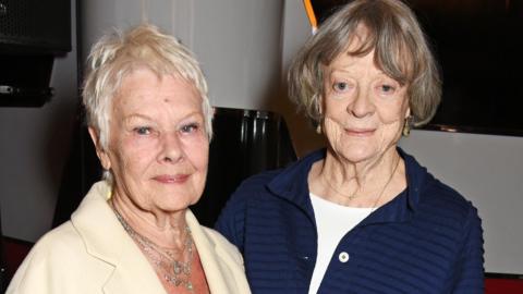 Dame Judi Dench, on the left of the image, a woman with cropped white hair wearing an ivory blazer and silver chains, and Dame Maggie Smith, a woman with dark grey hair in a bob wearing a navy jacket over a white top, both smiling