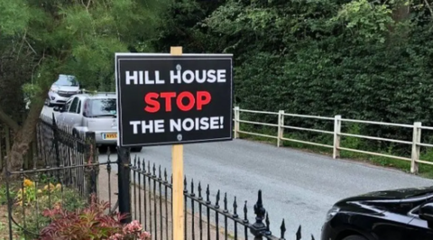 Black Placard on which is written Hill House in white capitals, STOP in red capitals and The Noise in white capitals, in a front garden beside a road with parked cars on it