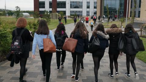 Nine teenage school pupils walk towards a school building at the start of the day. They are all wearing coats and carrying bags. They have their backs to the camera 