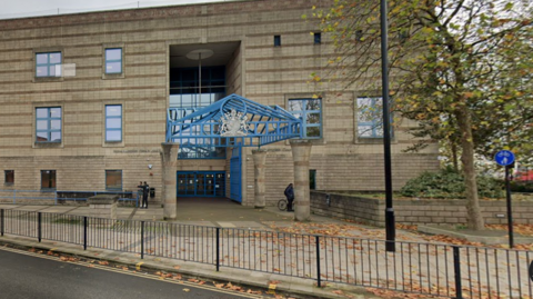 A google map view of the outside of a three-storey yellow-brick court building, with a blue metal canopy on pillars leading to blue doors at the entrance.