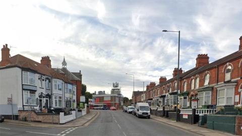 A Google Streetview picture of Issac's Hill in Cleethorpes showing terraced homes on either side of a residential street, with cars parked down the right-hand side and a large, two-storey building and bus in the background.