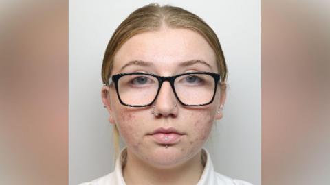 A woman with black-framed glasses, two nose piercings and a lip piercing looks at the camera.
