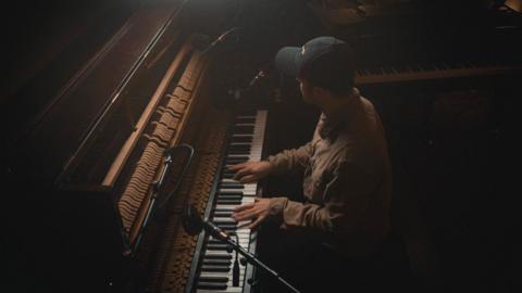 A man is wearing a black cap and a brown shirt. He is playing a piano in a darkly lit room. 