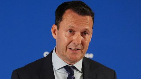 Russell Findlay wears a black blazer and grey shirt and is standing in front of a blue backdrop