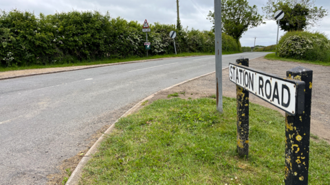 A sign which reads Station Road with a main road in the background with national speed limit signs