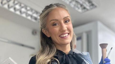 A woman with blonde hair is smiling at the camera. She is wearing a black top and a blue glove and standing in front of a light-coloured wall.