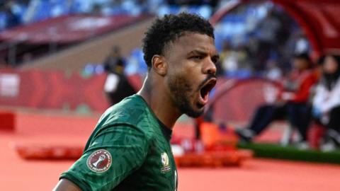 Lyle Foster visible from chest up opens his mouth wide to scream in delight after scoring a goal for South Africa against Zimbabwe at Afcon 2025. Foster is wearing a dark green jersey and is looking from left to right across the image. Blurred in the background is the orange running track which surrounds the pitch, with people sitting in a dugout