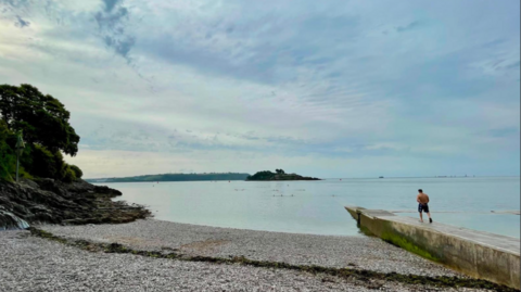 A calm seaside scene shows a pebbled beach and rocky shoreline leading to still water, with a small island in the distance and a person standing on a concrete jetty looking out to sea under a cloudy sky.