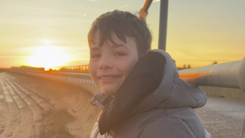 Lachlan is sitting outside in front of an orange sunset. He is facing sideways, smiling to the camera, and is wearing a grey hooded coat. He has short brown hair.