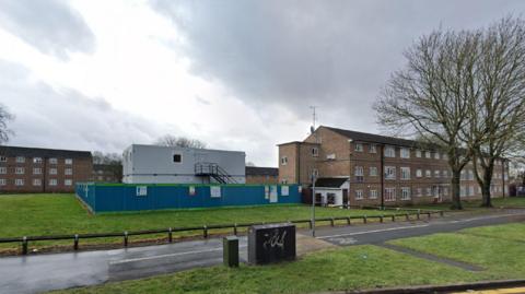 Brown brick blocks of flats surround a green space, on which sits a grey temporary building, surrounded by blue fencing.