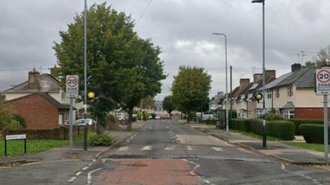 Image of a tree lined street with terraced housing on either side of the road. In the foreground can be seen a zebra crossing.