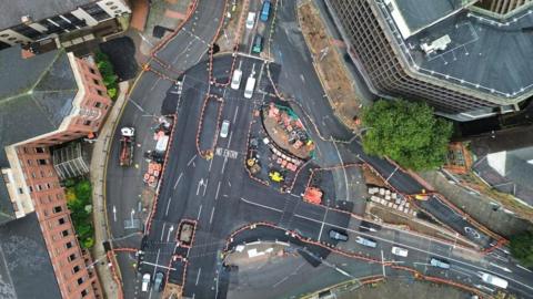 Drone shot of the junction, taken from directly overhead, showing new areas of road surface with some white lines and lots of construction barriers still in place