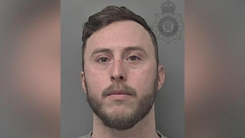 A police custody photograph of a man with short brown hair, a beard and stubble looking into the camera with a serious expression against a grey background.