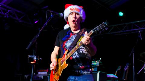 A man in a Santa hat with 'Slade' written on the white cuff in red lettering, plays the guitar as he looks into the camera while singing in front of a microphone on stage.