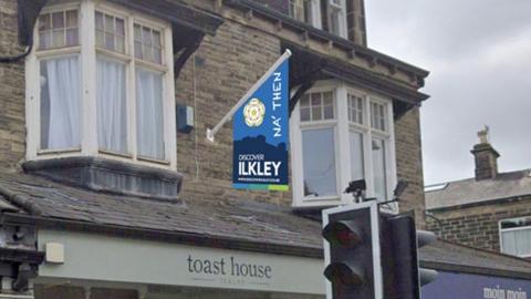 Row of shops on the ground floor and on the first floor a short flag pole juts out and from it hangs a flag in shades of blue showing the Yorkshire rose, the words Na Then and promotion for the town of Ilkley