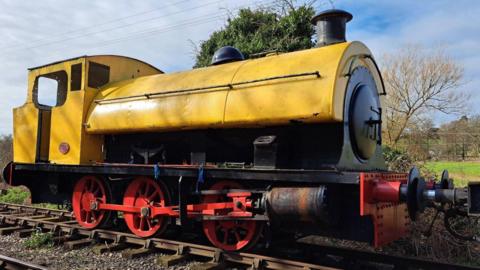 A steam locomotive, mainly yellow in colour, with red wheels, and resting on railway tracks. Trees are behind it. It has a cabin, and main carriage. 