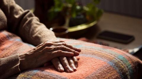 A senior person's hands folded across their lap onto a red thick blanket. They are sitting down and you can see a side table next to them and they are wearing a brown jumper.