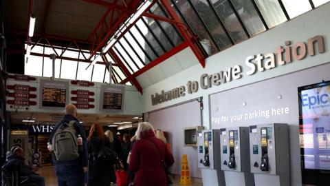 A view inside Crewe Station. Passengers are looking at information boards. There is a sign saying Welcome to Crewe Station.