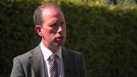 A clean-shaven man with short blonde hair wearing a grey suit and a stripy tie, photographed mid-talking on a sunny day with a hedge in the background.