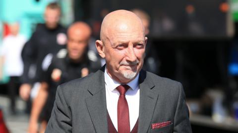 Ian Holloway wearing a grey suit, white shirt, red tie and waistcoat walking off the bus to the stadium at Swindon Town v Grimsby