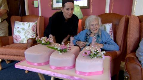 Centenaria Hilda Leggett sitting in an amrchair in front of a pink birthday cake in the shape of the numbers 110