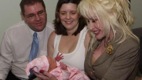 Dolly Parton holds a tiny baby in pink clothes while the baby's parents look on