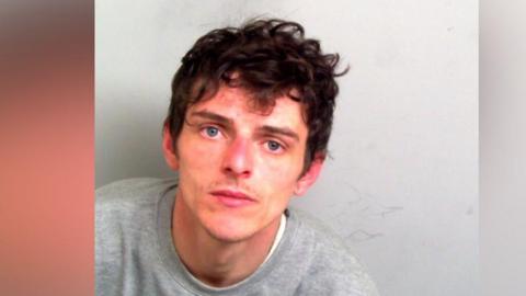 Dominic Boultwood in a police mugshot. He is wearing a grey sweatshirt and has dark hair and blue eyes.