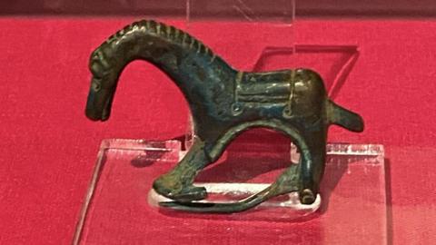 A small copper-alloy horse figurine which has a long curved neck is propped up on a clear stand on a red table.
