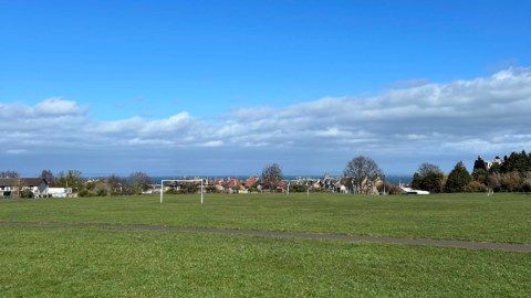 Wide open grassy field at Joppa Quarry Park with two white football goals, a paved path in the foreground, houses and trees in the distance, and a blue sky with scattered clouds above.