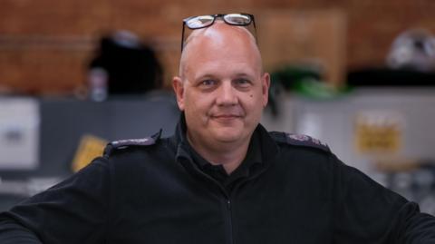 A bald man with glasses on his head. He is wearing a black jumper.