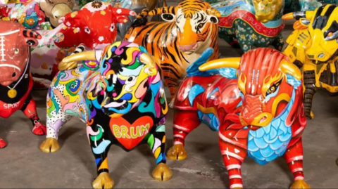 Close-up photos of a group of nearly life-size fibre-glass statues of bulls, brightly painted in various designs. The one in the foreground at the front of the picture has a red heart painted on its chest with the letters BRUM in gold inside. Another next to it is painted red and blue to look like a chinese Dragon.

