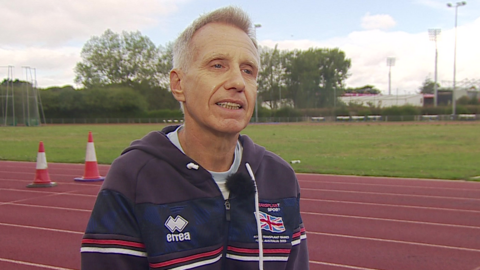 Vince Taylor is sat answering questions next to a racetrack and he is wearing a blue hooded top with the Great Britain flag. He has short, white hair.