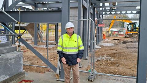 Man in white hard hat and high-visibility jacket stands on muddy ground at a construction site, facing a steel framework structure with beams and columns; excavator and building materials are visible, with a "Do Not Enter" sign on a staircase and temporary fencing surrounding the area.