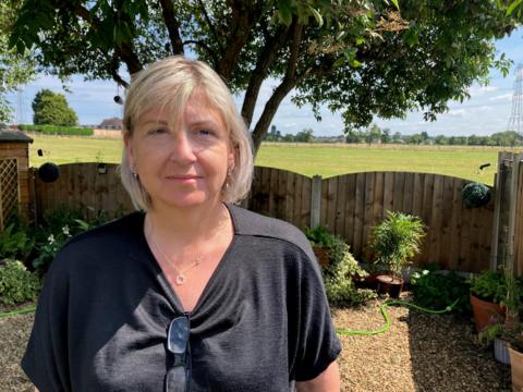 A woman with chin-length blonde-grey hair in a bob stands in a garden on a sunny day. A fence behind her looks out over a large field.