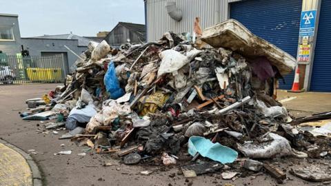 A large pile of rubbish that has been fly-tipped on an industrial estate outside a garage. There is bedding, mattresses as well as wood in the dump.