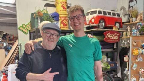 Two men in glasses, each with an arm around the shoulder of the other. They are both smiling, the older man is wearing a flat cap