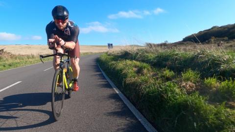 A man with a cycling helmet and glasses rides a green racing road bike on a road with fields either side. 