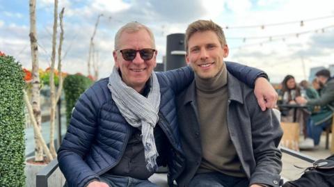 The image shows two men sitting next to each other on an outdoor bench. One person has an arm resting around the shoulders of the other, and both are facing the camera. The person on the left is dressed in a dark quilted jacket, a light scarf, dark trousers, and sunglasses. The person on the right is wearing a dark coat over a high‑neck jumper and dark trousers. 