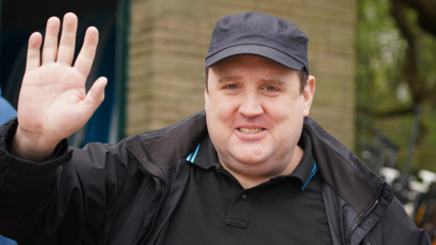 A man with short dark hair, a dark coat, dark polo shirt and dark cap with one hand raised, palm forward